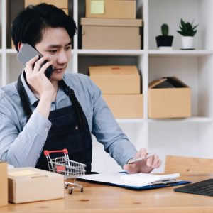 Young asia seller man happy work on laptop busy cellphone call chat to customer at home office desk