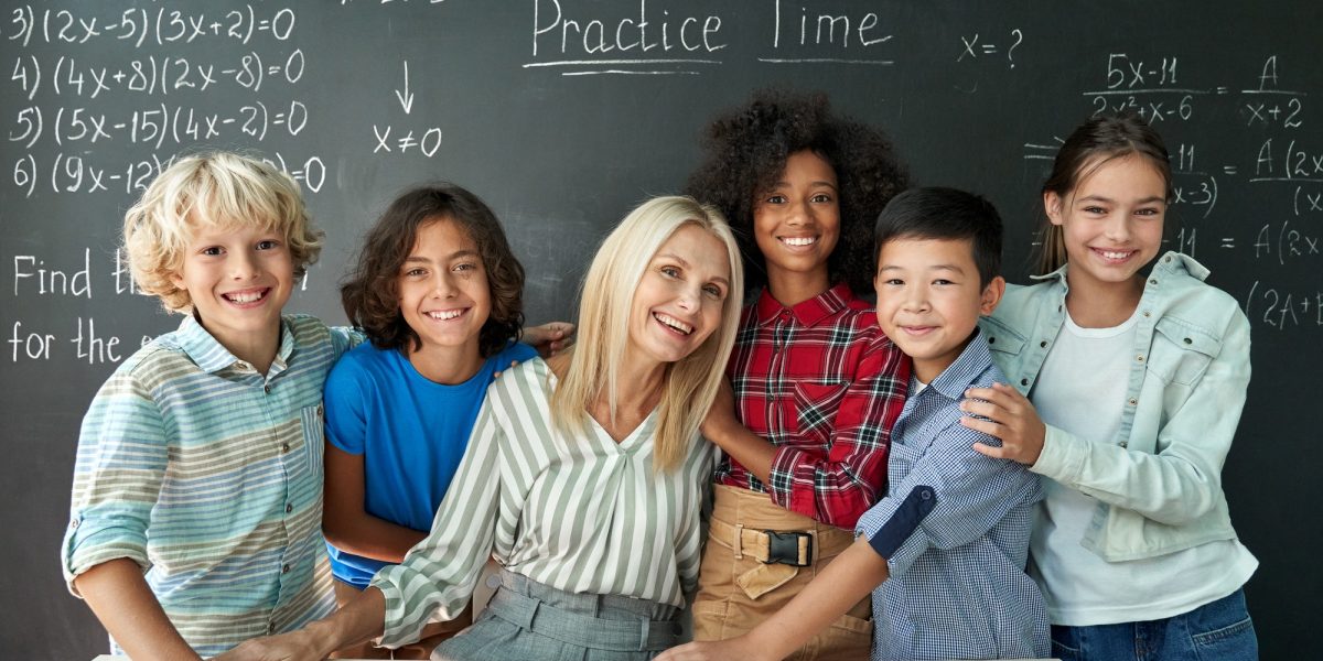 Portrait of cheerful smiling teacher and diverse schoolkids near chalkboard.
